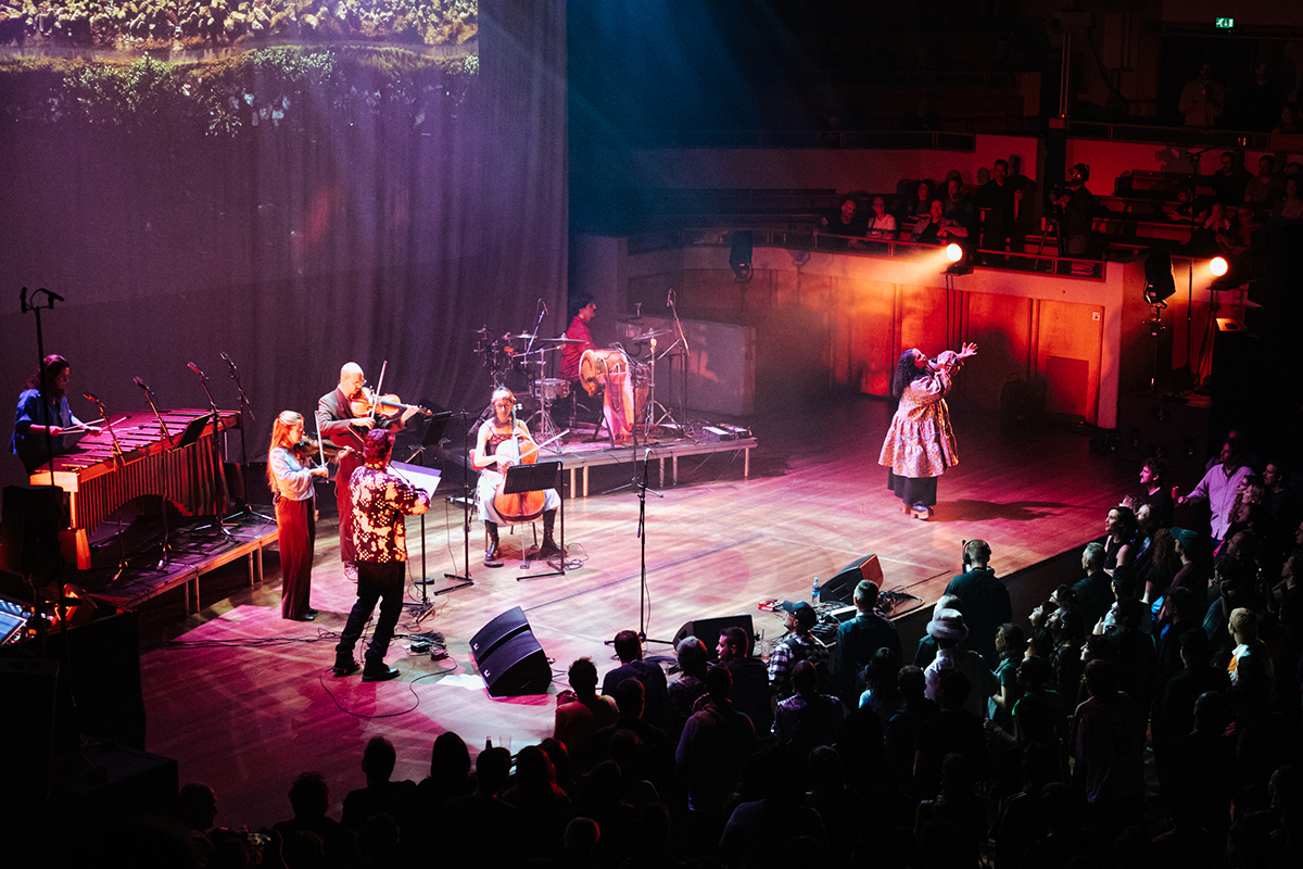 Lido Pimienta singing on stage at Le Guess Who? with one hand raised in the air. On stage is also a string quartet, marimba player and percussionist